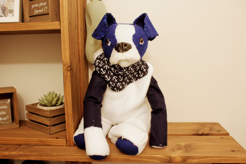 Stuffed toy dog with a purple bow and anchor-patterned bandana on a plain background